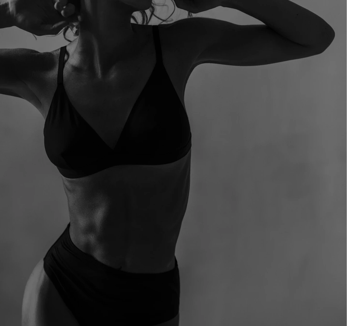 Woman in black bikini posing against neutral background, showcasing torso and body contour